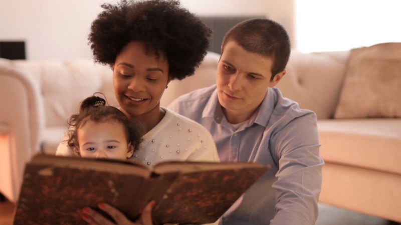 Two parents and a child sitting near a beige couch, reading a large book together.